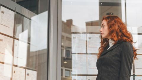 Young woman in a suit with long curly hair looking at job adverts in a window.