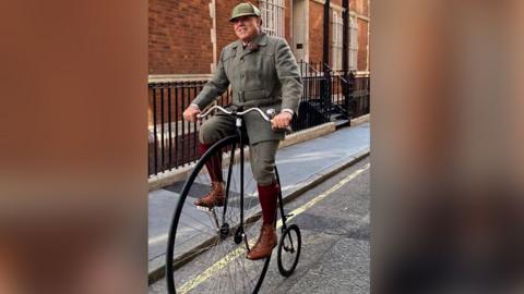 Brian Cobbold is wearing a tweed outfit including a hat and brown boots. He is on a street riding a penny farthing cycle, which has a huge front wheel and a small back one.