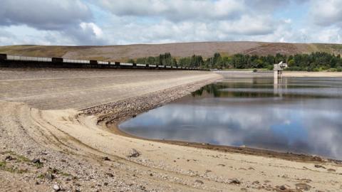 Backwater Reservoir, Angus. The water is well below the level of the wall on the left of the image, and there is sand in the foreground and to the left.