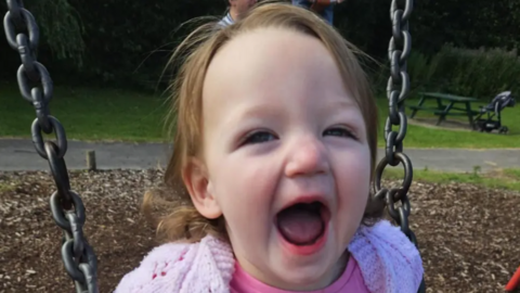 A head and shoulders picture of a two-year-old girl, Lyla, while on a swing in a children's park. She has short brown hair and is smiling broadly at the camera. She is wearing a pink cardigan and pink top.