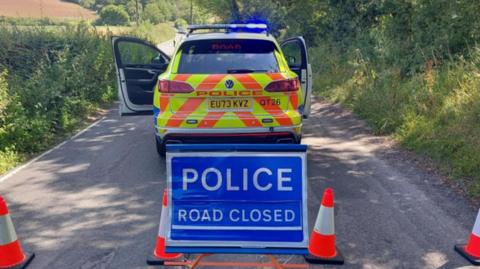 A blue sign that reads "Police Road Closed" with orange cones to its right and left, with a police car behind it blocking the lane.