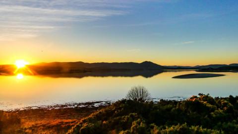 Sun setting behind hills with a still body of water in the foreground