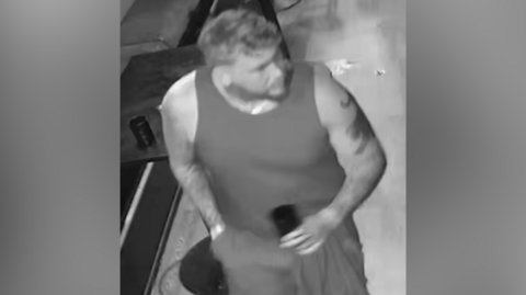 A man in a dark vest and casual bottoms with short hair and a tattoo on his arm. He is standing on a wooden floor. 