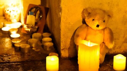 Candles, a teddy bear, and a holy water font lie on the ground.