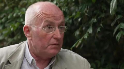 Spt Gerry Murray sitting before a bush wearing a white shirt, a beige jacket and glasses