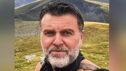 Chris Ellery standing on a green mossy mountain top. He has a grey beard