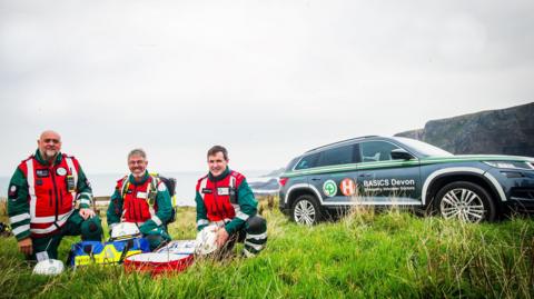 Three men in red and green hi-vis jackets and black hi-vis trousers, kneeling around medical equipment with a car to the side of them with the words 'BASICS Devon' printed on the side.