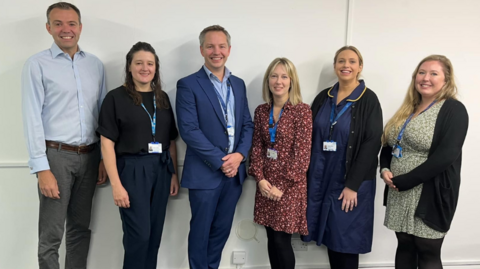 The team working on the project at Somerset NHS Foundation Trust - two men and four women are standing against a white wall, smiling.
