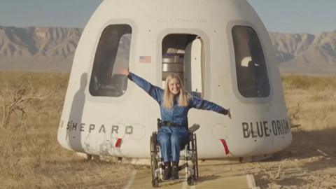 woman in blue boiler suit in wheelchair sits with arms out wide in front of Blue Origin capsule in a desert with mountains seen in the far distance