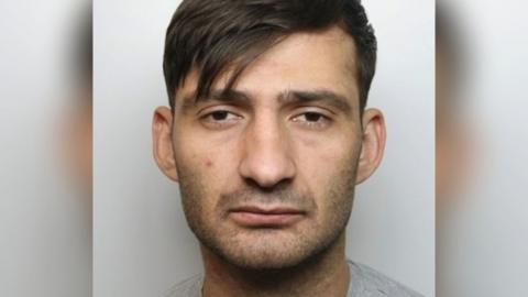 A police mugshot of Sulejman Zekthi. He has stubble around his lower face and has brown hair swept to one side in a fringe. He is wearing a greay t-shirt and is standing against a white background.