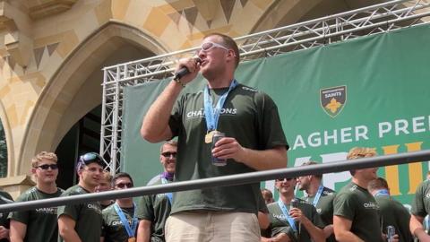 A man wearing a green sports jersey and a medal is holding a microphone. His team mates are stood behind him in front of a large green stage background