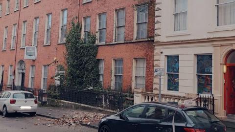 A street photo showing a row of terraced Georgian-style buildings.  A pile of bricks lie on the pavement below the building in the middle of the photo.  Cars are parked on either side of the bricks. 