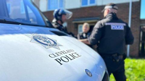 Library image of a Cleveland Police van. The force's logo can be seen on the bonnet of the silver vehicle. Three officers wearing uniforms are talking to each other in the background.