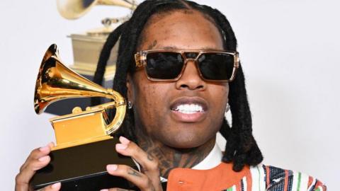 Lil Durk wearing sunglasses and a multi-coloured suit, and holding up the gold gramophone-shaped Grammy Award