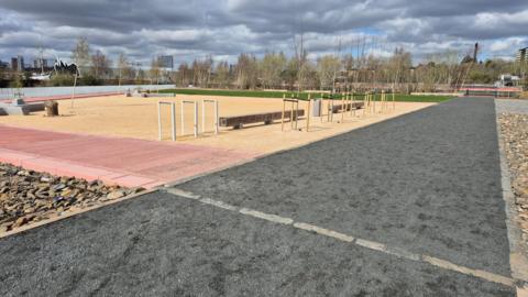 An outdoor park, with with greenspace, stones, a sandy area in the middle and a path with ash on it.