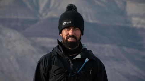 A man with a dark brown beard and wearing a black woolly hat and jacket is standing looking directly into the camera. He is smiling with mountains in the background.