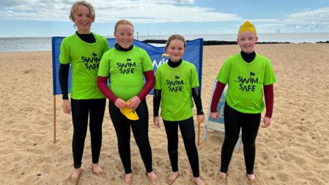 Four children wearing hi-vis T-shirts and wetsuits stand on a beach and smile at the camera.