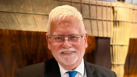Nick Allan has white hair and a short white beard. He is wearing a black suit and a blue tie. He has glasses and is standing in front of an Angel of the North statue. 