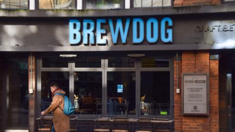A person walks past a Brewdog bar. The sign on the wall has the name of the company in large blue letters.