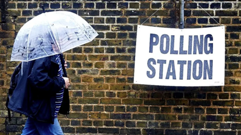 a person holding an umbrella over their head walks past a sign hanging from a wall which says Poling Station