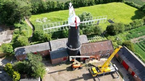 An aerial picture of two sails placed horizontally on a windmill. The sails are white and they are placed on a white windmill cap. The windmill is in a courtyard surrounded by farm buildings and green fields.