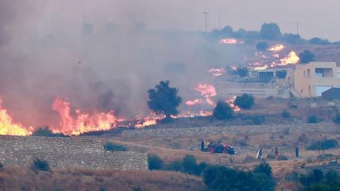A fire burns through low-lying vegetation across an arid slope towards a village as people watch from nearby.