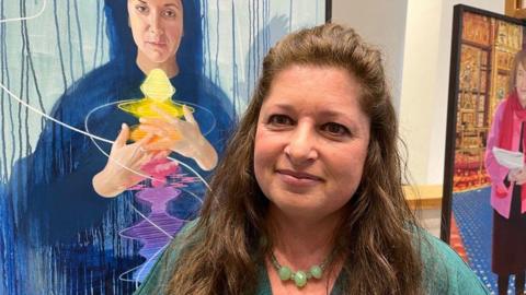 A woman in a green cardigan and necklace stands in front of some painted portraits of women.