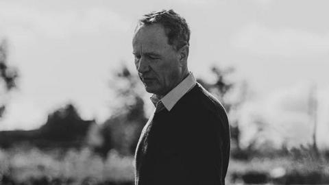 A black and white photo of David Austin in the middle of a field of flowers, looking down at them. He is wearing dark trousers with a black jumper and a white collar