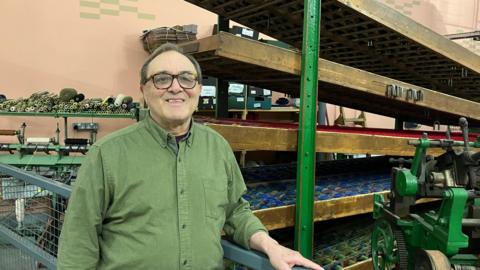 A man wearing a green shirt and glasses stands in front of a large wooden loom with a pink wall behind him.
