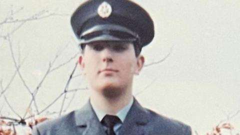 A man is dressed in a RAF uniform. He is stood in front of a hedge which is orange in the autumn. The sky is grey