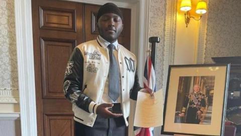 Young man wearing varsity-style jacket, shirt and tie - he stands next to a picture of the king and a British flag.