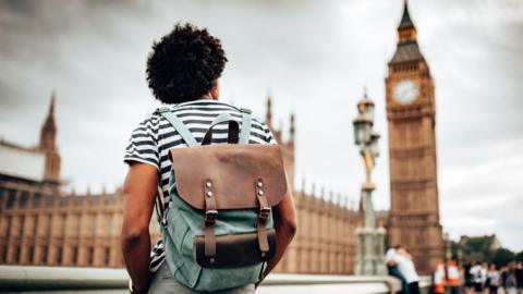 A man with dark brown afro hair standing with his back to the camera wearing a black and white horizontal striped t-shirt and a brown and green backpack looking up at the Houses of Parliament