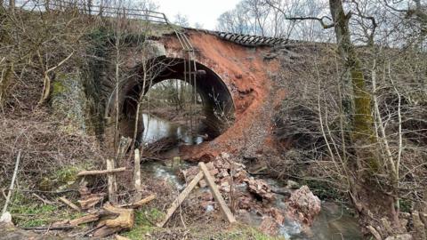 A bridge underneath a railway with a river running underneath it. Earth has fallen away from the railway track leaving it exposed. There is debris and fencing in the river below.