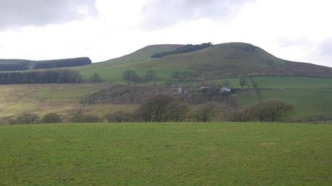 The site of a British hillfort near Lockerbie - a large mound of earth, completely covered with grass set in rural countryside with trees, fields and what looks like a farm building
