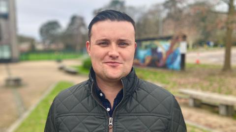 Dark-haired man posing for a photo with trees and benches behind him, wearing a black jacket 