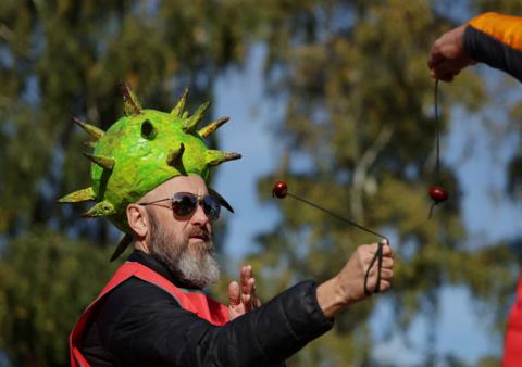 A bearded man wearing a horse chestnut shell model hat on his head. He is playing conkers with a conker on the end of a black string. 