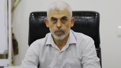 A headshot of Yahya Sinwar, wearing a white dress shirt, sitting in a black leather desk chair and staring into the camera