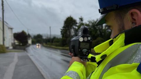 A man in a high visibility police jacket and cap points a hand-held speed gun at a wet road.