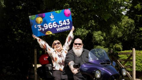 A woman holding a sign that says "The National Lottery and £3,966,543 winner". The background of the sign in clue and there multicoloured bingo balls. There is a man sat in front of the woman on a purple Harley-Davidson trike bike. There are trees behind them and a wooden fence.  