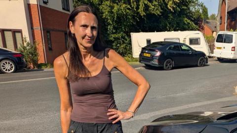 Zoe Farmer stood beside her black car which has scratches and dents along the front. She has brown hair and is wearing a brown top with black shorts.