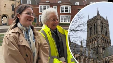 Female reporter, with brunette hair, glasses and a beige trench coat is stood next to a tour guide of Lincoln Cathedral. She is an older lady wearing a yellow hi-vis jacket. Both are smiling past camera and looking up.