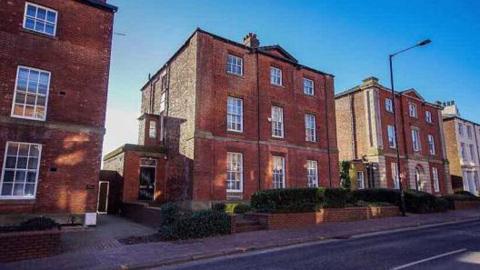 A red-brick three-storey building stands on a residential street, behind a garden wall and a small hedgerow