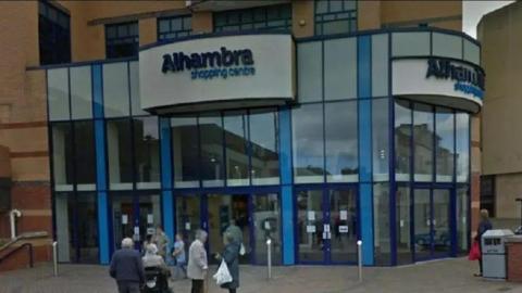 The front of the Alhambra Shopping Centre with a group of about nine people standing in front of it on a brick concourse