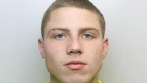 Police mugshot of a man with a wide face but pointy head and chin. He has thin long eyebrows and blue eyes..