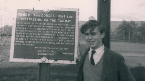 Black and white photo of Paul Antell as a teenager standing next to a sign which explains it is forbidden to trespass on the Somerset and Dorset line.