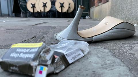 A discarded high-heeled shoe lies on a pavement next to an empty cigarette pack and small debris. In the background, outdoor tables are folded up outside a venue.