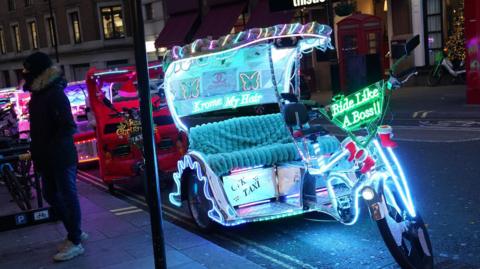 A brightly illuminated pedicab on a London street at night, adorned with colorful neon lights, festive decorations, and playful text like "Ride Like A Boss!" Nearby, a person in winter clothing stands next to bicycles, with surrounding shops and signage in the background.