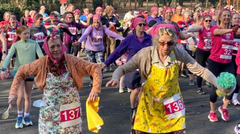 A large crowd of people, the vast majority women, warm up for a four-mile run. Many are wearing pink headscarves. At the front, two women are dressed in vintage headscarves, aprons and cardigans with competitor numbers pinned to the clothes.