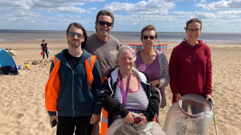 Two men and three women stand with refuse bags and litter pickers at Sutton on Sea beach, they are wearing hi-vis vests. Families can be seen enjoying a day out in the background.