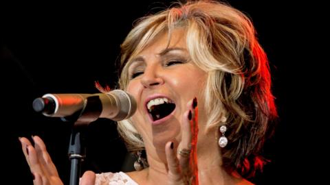 Lesley Garrett, singing, with her arms help up, she has her mouth open and has earrings in her ears. She has short fair hair. 